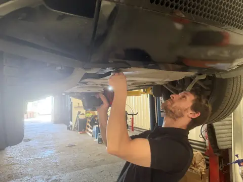 Russ under a car raised on the ramp, using an impact gun on the underside at RPM Autocare, Newton Abbot